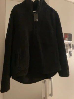 Svart teddyjacka med dragkedja - Mysig svart teddyjacka från Lager 157 med hög krage och dragkedja framtill. Jackan har en avslappnad passform och är tillverkad i fluffigt teddy-material som håller dig varm. Perfekt för kalla dagar och enkel att matcha med olika outfits.