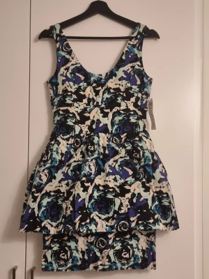 Klänning Lindex - Säljer en snygg kortklänning med blommigt mönster i blå, svart, vit och beige. Klänningen har breda axelband, markerad midja och en voluminös kjoldel som ger en cool siluett. Perfekt för dig som vill sticka ut med färg och mönster!