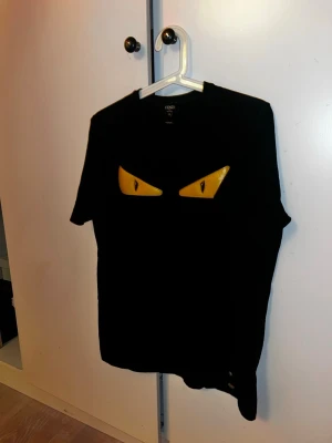 Svart Fendi t-shirt med gula ögon - Cool svart t-shirt från Fendi med ikoniska gula ögon på bröstet. Klassisk passform och rund halsringning. Perfekt för dig som vill sticka ut med en lyxig och unik detalj. Fendi-logga på ärmen.