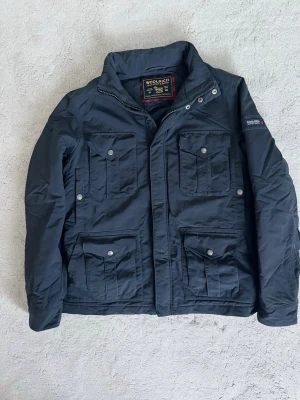 Svart parkajacka från Woolrich - Snygg parkajacka från Woolrich med fyra stora fickor framtill och diskret logga på ärmen. Jackan har hög krage, dragkedja och tryckknappar. Tillverkad i slitstark polyester och passar perfekt till kyligare dagar. Modellen är ca 182, 70 kg.