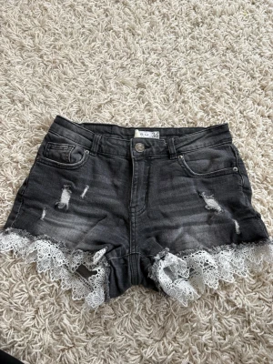 Grå denimshorts med spetsdetalj - Snygga grå denimshorts från S.U. i storlek 36. De har hög midja, slitna detaljer och vit spets längs bensluten som ger en cool och unik look. Perfekta för sommarens alla äventyr och festivaler.
