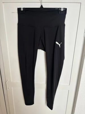 Svarta leggings från Puma - Snygga svarta leggings från Puma med hög midja och klassisk vit logga på benet. Tillverkade i stretchigt och mjukt material som sitter skönt på kroppen. Perfekta för träning eller chill dagar.