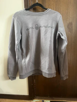 Grå Sail Racing sweatshirt - Snygg grå sweatshirt från Sail Racing med tryckt logga på bröstet och en mindre logga i nacken. Tröjan har rund halsringning, långa ärmar och ribbade muddar. Säljer för att den är för liten. Storlek 160.