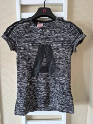 Gråmelerad Adidas t-shirt med A-print - Cool gråmelerad t-shirt från Adidas med stort svart A-print på bröstet. T-shirten har korta ärmar med snygga öppna detaljer på axlarna och är tillverkad i mjuk polyester. Perfekt för dig som gillar sportig stil och vill sticka ut.