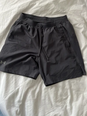 Grå träningsshorts från Under Armour - Säljer ett par träningsshorts från Under Armour med snörning i midjan och diskret logga på benet. Shortsen har sidofickor och är tillverkade i ett lätt och snabbtorkande syntetmaterial, perfekta för gymmet eller löprundan.