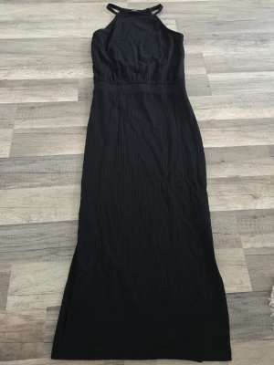 Svart långklänning med halterneck - Stilren svart långklänning med halterneck och smala axelband. Klänningen har en rak passform och är ärmlös, vilket ger en elegant siluett. Perfekt för dig som gillar minimalistisk och tidlös stil.