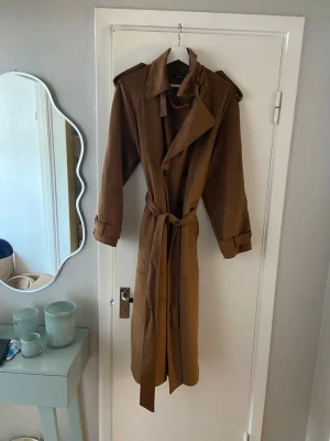 Brun lång trenchcoat från Gina Tricot - Stilren brun trenchcoat från Gina Tricot med klassisk krage, axelklaffar och knytskärp i midjan. Jackan har långa ärmar och är tillverkad i ett mjukt, mockaliknande material. Lätt fodrad . Jättefin. Stl M
