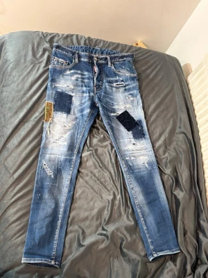 Dsquared jeans - Säljer ett par blå jeans med skinny fit och coola slitningar, patchar i olika färger och detaljer som ger en unik streetwear-look. Jeansen har flera lagade hål och färgstänk för en trendig vibe. Perfekta för dig som gillar en edgy stil.