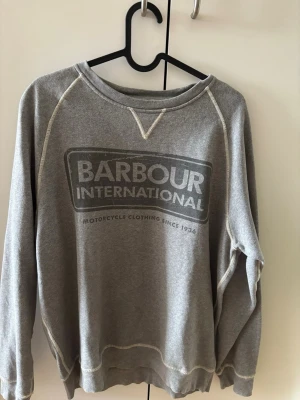 Grå sweatshirt från Barbour - Säljer en grå sweatshirt från Barbour International med tryck på bröstet och kontrastsömmar. Tröjan har rund halsringning och långa ärmar. Perfekt för en avslappnad och stilren look. Materialet känns mjukt och bekvämt.