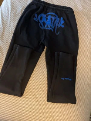 OG syna world sweatpants - Säljer ett par helt oanvända svarta mjukisbyxor med blått SYNA WORLD tryck på baksidan. Byxorna är gjorda i mjukt o myxket skönt  material, perfekta för streetwear. Snygg streetwear vibe! Äkta såklart då de är köpta från Strtsply