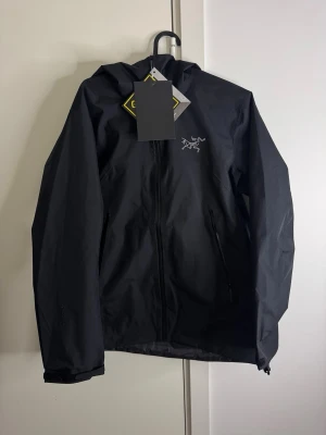 Svart vindjacka från Arc'teryx - Snygg svart vindjacka från Arc'teryx med dragkedja framtill och två sidofickor med dragkedja. Jackan har huva och diskret logga på bröstet. Tillverkad i ett lätt och slitstarkt material som passar perfekt för blåsiga dagar.