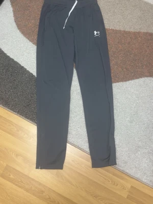 Svarta träningsbyxor Under Armour - Snygga svarta träningsbyxor från Under Armour med vitt logotryck på låret och dragsko i midjan. Byxorna har raka ben och sidofickor, perfekta för gymmet eller löprundan.