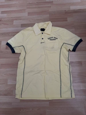 Ny. Gul piké Sunset Diner Glendale - Gul pikétröja med svart text 'Sunset Diner Glendale' på bröstet. Tröjan har krage, korta ärmar med mörka muddar och knappslå framtill. Dekorativa svarta sömmar längs sidorna och en bröstficka. Perfekt för en avslappnad stil.