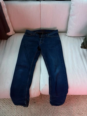 Mörkblå Nudie jeans - Mörkblå nudie jeans med storlek 32/32. Guldfärgade sömmar. Sitter inte skinny utan snarare vanligt. Kan skicka bild om det önskas. Gratis reparation hos Nudie om de går sönder.