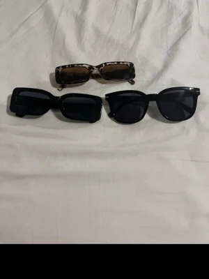 Tre trendiga solglasögon i olika former - Säljer tre par solglasögon: ett par med leopardmönstrad båge och bruna glas, ett par svarta med rektangulär form och ett par svarta med klassisk rundad båge. Perfekta accessoarer för att lyfta din stil i solen.