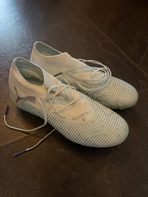 Ljusblåa fotbollsskor från Puma - Säljer ett par stilrena vita fotbollsskor från Puma med ljusblå detaljer och ribbad ovansida för bättre bollkontroll. Skorna har snörning och en strumpliknande krage för skön passform. Perfekta för dig som vill sticka ut på planen.