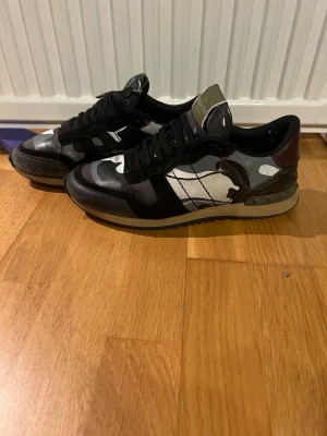 Valentino rockrunners  svart/grå camo  - ÄKTA Snygga sneakers från Valentino med svart och grått camo-mönster, svarta skosnören och detaljer i mörkrött vid hälen. Skorna har en låg silhuett och är tillverkade i en mix av läder och textil. Perfekta för dig som vill sticka ut med en exklusiv touch.