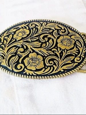 Ovalt bältespänne med blommigt mönster - Snyggt ovalt bältesspänne i metall med detaljerat blommigt mönster i guldton. Perfekt för att ge din outfit en unik touch. Passar till de flesta bälten och är ett riktigt statement piece för dig som gillar accessoarer med karaktär.