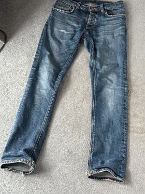 Grim Tim Nudie Jeans 30/32 - Säljer ett par klassiska blå jeans från Nudie Jeans med slitningar och snygg tvätt. Jeansen har raka ben och detaljerade sömmar på bakfickorna. Materialet är denim i bomull och passformen är relaxed fit. Perfekta för en avslappnad stil. Pris går att diskutera. 