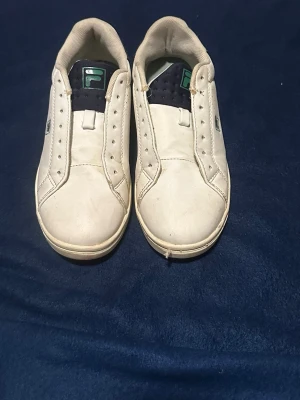 Vita Fila sneakers med gröna detaljer - Klassiska vita sneakers från Fila med gröna och mörkblå detaljer. Skorna har lågt skaft, perforerade sidor och Fila-logga på sidan och plösen. Ovandelen är i läder eller syntet och sulan är vit. Perfekta för en clean och sportig look. Snören medföljer 