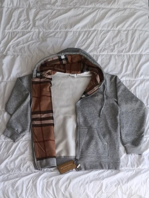 Grå zip hoodie från Burberry - Snygg grå zip hoodie med klassiskt rutigt foder i huvan. Hoodien har dragsko i huvan, fickor framtill och ribbade muddar. Materialet är mjukt och bekvämt, perfekt för en chill och stilren look.