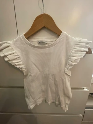 Vit ribbad topp med volangärmar Zara - Supersöt vit ribbad topp från Zara med bred rund halsringning och volangdetaljer på ärmarna. Perfekt att styla till jeans eller kjol för en fräsch look. Materialet känns mjukt och stretchigt.