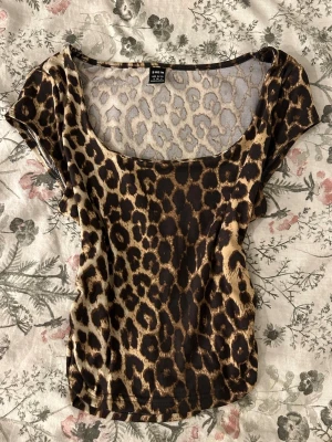 Leopardmönstrad topp från SHEIN - Trendig topp från SHEIN med leopardmönster i bruna och svarta toner. Modellen har korta ärmar, rundad urringning och är figurnära. Perfekt för dig som vill sticka ut med ett djuriskt print.