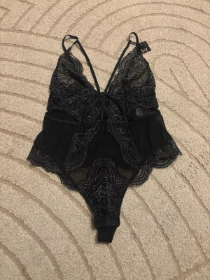 Svart spetsbody med tunna axelband - Snygg svart body i spets från Hunkemöller med tunna axelband och djup urringning. Bodyn har transparent spets med blommönster och dekorativ rosett framtill. Perfekt för dig som gillar feminina detaljer och en elegant look.