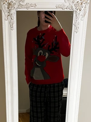 Röd jultröja med renmotiv och lampor - Rolig röd jultröja med stort renmotiv framtill och inbyggda blinkande lampor i hornen. Tröjan har rund hals och långa ärmar, perfekt för julens alla tillfällen. Materialet är mjukt och bekvämt, passar dig som vill sticka ut på julfesten.