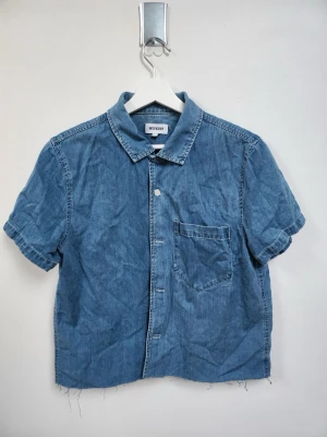 Blå kortärmad skjorta från Weekday - Säljer en blå kortärmad skjorta från Weekday i denim-look med klassisk krage och bröstficka. Skjortan har knappar framtill och rå, fransig nederkant för en cool vibe. Perfekt för dig som gillar avslappnad stil.