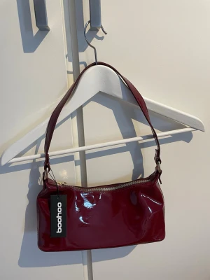 Vinröd axelväska från Boohoo - Snygg vinröd handväska från Boohoo i glansigt material med enkel axelrem och dragkedja upptill. Nypris 270kr