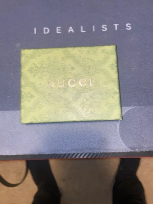 Gucci plånbok med GG-mönster - Snygg plånbok från Gucci med klassiskt GG-mönster i beige och brunt. Plånboken har gröna och röda ränder på framsidan och levereras i en elegant grön ask med Gucci-logga. Perfekt för dig som gillar exklusiva accessoarer.