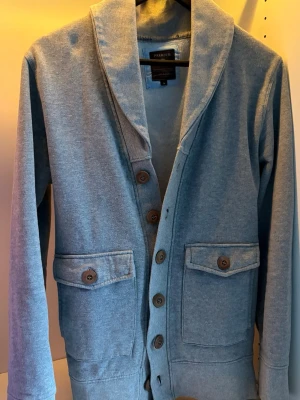 Cardigan Jack & Jones Premium  - Stilren ljusgrå kofta från Jack & Jones Premium med stora bruna knappar och två fickor framtill med lock. Koftan har en bred sjalkrage och är i  bomullsmaterial.
