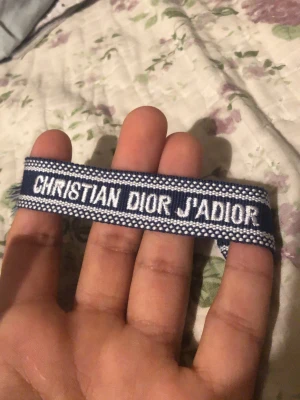 Blått Christian Dior J'Adior armband - Snyggt blått tygarmband från Christian Dior med texten 'J'ADIOR' broderad i vitt. Armbandet har en klassisk och trendig look med vita detaljer längs kanterna. Perfekt accessoar för dig som vill ha något stilrent och ikoniskt från Dior.