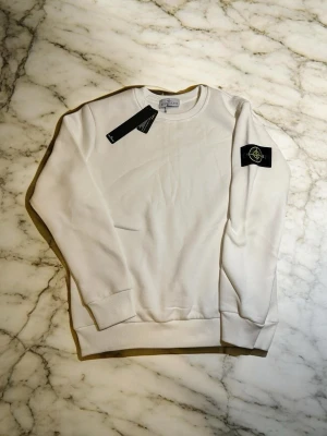 Vit sweatshirt från Stone Island - Säljer en clean vit sweatshirt från Stone Island med den ikoniska svarta patchen med gult och grönt märke på vänster ärm. Tröjan har rund hals, ribbade muddar och är gjord i mjukt bomullsmaterial. Perfekt för dig som gillar streetwear och vill ha något stilrent.