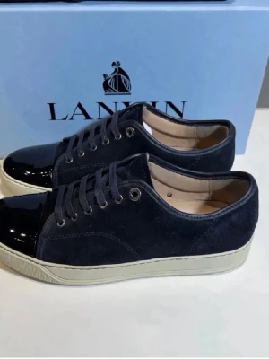 Lanvin svarta låga sneakers mocka - Snygga svarta sneakers från Lanvin med ovandel i mocka och lackad tå. Skorna har vita gummisulor och svarta skosnören. Klassisk låg modell med metallöljetter och diskret logga på sulan. Perfekta för dig som gillar stilrena och exklusiva sneakers.