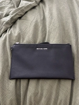 Svart kuvertväska Michael Kors - Stilren svart kuvertväska från Michael Kors i präglat läder med silverfärgad dragkedja och diskret logga framtill. Väskan har en avtagbar handledsrem och minimalistisk design som gör den enkel att matcha med olika outfits.