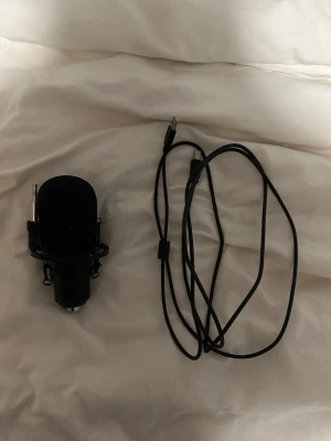 Svart USB-mikrofon med kabel - Säljer en svart USB-mikrofon med puffskydd och tillhörande USB-kabel. Mikrofonen har en rundad form och sitter i ett stötdämpande fäste. Perfekt för streaming, podd eller gaming. Enkel att koppla till datorn via USB.