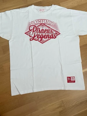 Olympiacos Piraeus Legends vit t-shirt XL - Vit t-shirt från Olympiacos med röd tryckt text 'Piraeus Legends' på bröstet och klubbmärke på ryggen. Klassisk passform med korta ärmar och rund hals. Materialet är mjuk bomull och t-shirten har en patch längst ner på framsidan.