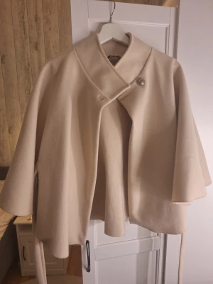 Beige cape-kappa med knytband - Säljer en beige cape-kappa med knytband i midjan och stora, vida ärmar. Kappan har en enkel knapp framtill och är tillverkad i ett mjukt tyg som ger ett snyggt fall. Perfekt för dig som gillar stilrena och trendiga plagg.
