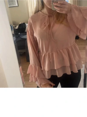 Rosa volangblus med knyt från H&M - Supersöt ljusrosa blus från H&M med volanger både vid ärmslut och midja. Blusen har långa ärmar och ett knytband vid halsen för extra detalj. Tillverkad i ett tunt, flowy material som ger en romantisk vibe. Är i storlek XXS men passar mig som har Xs-M. Har en liten fläck på ärmen - men utöver de inga defekter.