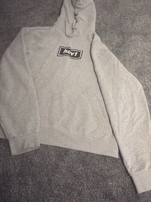 Grå hoodie från nov1 med tryck - Snygg grå hoodie från nov1 med svartvitt tryck på bröstet. Klassisk känguruficka framtill, ribbade muddar och justerbar huva med dragsko. Perfekt för en chill och avslappnad stil. Tillverkad i mjukt sweatshirtmaterial.