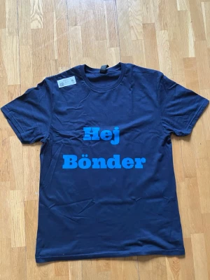 Blå t-shirt med DIF-tryck - Mörkblå t-shirt med stora blå texter på både fram- och baksida. På framsidan står det 'Hej Bönder' och på baksidan 'Vi é DIF!'. Klassisk rund hals och korta ärmar. Perfekt för dig som vill visa ditt stöd för Djurgården.