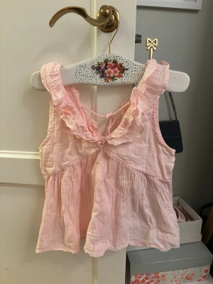 Ljusrosa volang topp med knyt - Supersöt ljusrosa linnetopp med volangdetaljer runt ärmhålen och knyt i ryggen. Luftig och flowy modell som passar perfekt till sommaren. Tillverkad i mjukt bomullstyg för en bekväm känsla.