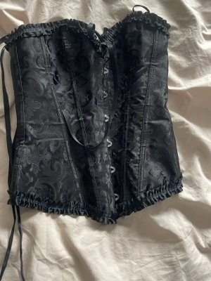 Svart mönstrad korsett med snörning - Snygg svart korsett med blommigt mönster och dekorativa volangkanter. Snörning fram och bak för justerbar passform. Tillverkad i glansigt tyg som ger en lyxig känsla. Perfekt för att skapa en elegant siluett.