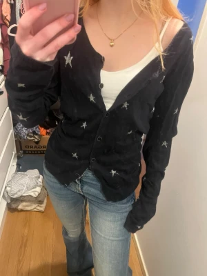 Mörkblå kofta med stjärnor - Säljer en svart kofta med silverfärgade stjärnor broderade över hela plagget. Koftan har knäppning framtill och långa ärmar. Perfekt för dig som vill ha en unik och trendig look med lite extra detaljer.