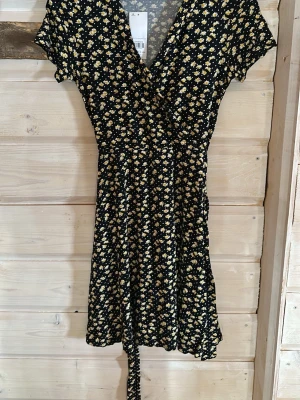Blommig omlottklänning  - Söt kort omlottklänning från Dorothy Perkins med svart botten och små gula blommor. Klänningen har v-ringning, knytband i midjan och korta ärmar. Perfekt för sommarens alla tillfällen och ger en fräsch och ungdomlig look.