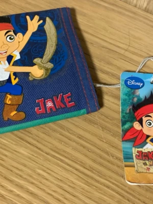 Jake och piraterna plånbok Disney - Blå plånbok med motiv från Jake och piraterna i Never Land. Plånboken har röda och gröna detaljer, tryckt text och illustration på framsidan samt flera fack inuti. Tillverkad i slitstark textil, perfekt för att hålla koll på kort och mynt.