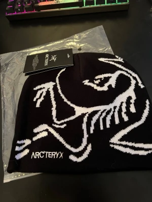 Svart mössa från Arc'teryx - Snygg svart mössa från Arc'teryx med stor vit logga och grafiskt skelettmönster. Tillverkad i mjukt, stretchigt material som sitter skönt på huvudet. Perfekt för dig som gillar streetwear och outdoor-vibe. Klassisk passform och stilren design.