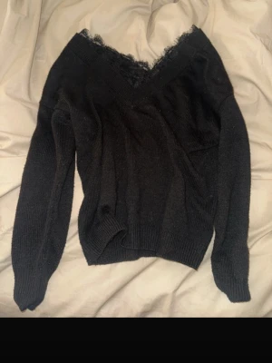 Svart stickad tröja med spetskant - Snygg svart stickad tröja med v-ringning och dekorativ spetskant vid halsen. Tröjan har långa ärmar och ribbade muddar vid ärmslut och nederkant. Perfekt för en trendig och stilren look.